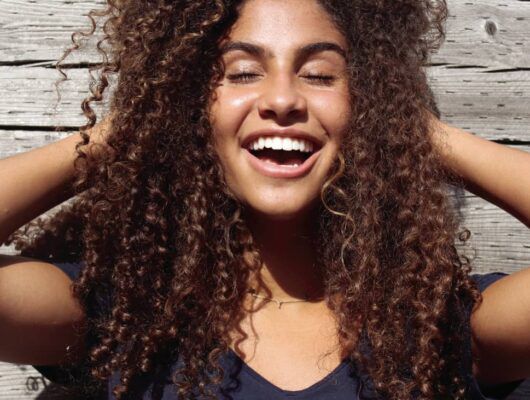 Woman with curly hair running her hands through her hair while smiling with her eyes closed in front of a wood background.